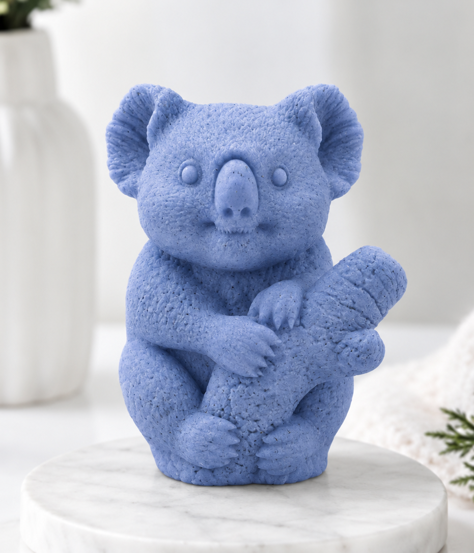 A front-facing photograph of a koala-shaped soap bar in a vibrant periwinkle indigo blue shade. The detailed soap is sculpted to show textured fur and the koala clinging to a log. It sits on a round, white marble disc. The blurred background shows a white vase with greenery and a rolled white towel