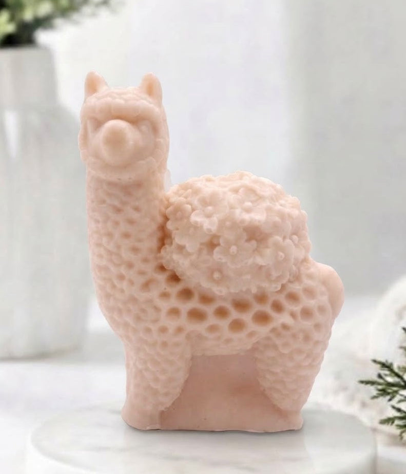 A product photo of a soft pink French clay llama / alpaca-shaped soap, standing on a white marble pedestal. The intricate carving details the texture of its curly fleece and the cluster of small flowers on its back, set against a softly blurred white bathroom background with green foliage and a white vase.