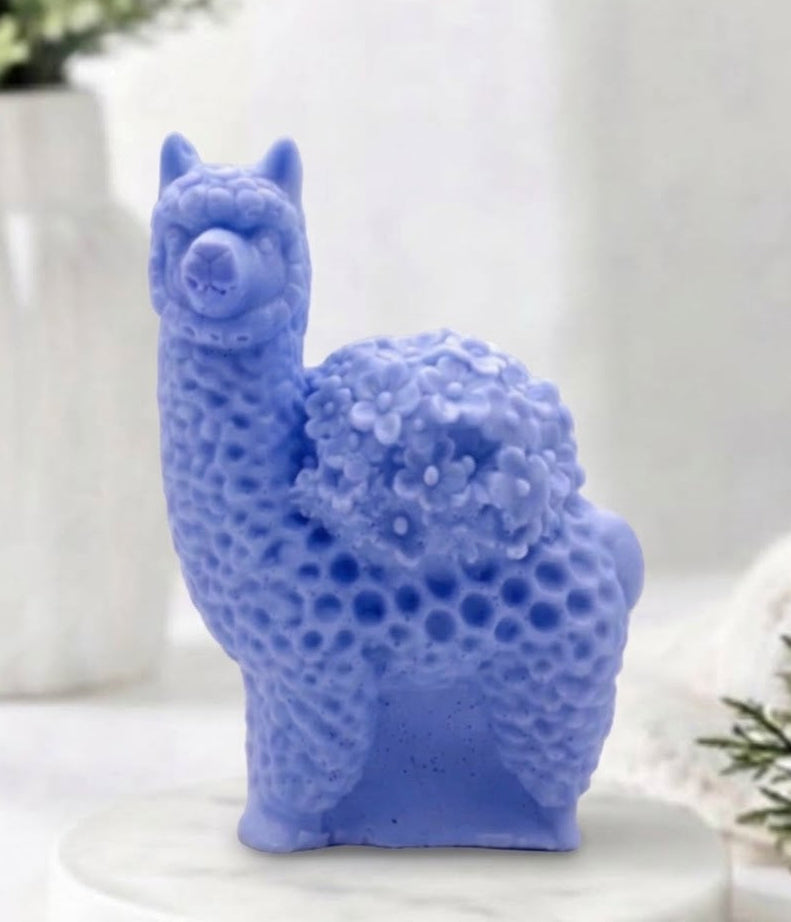 A product photo of a vibrant, indigo-blue llama / alpaca-shaped soap standing on a white marble disc, with a blurred white bathroom and eucalyptus plant in the background. The soap is highly detailed with a distinctive curly fleece texture and a bouquet of small flowers on its back.