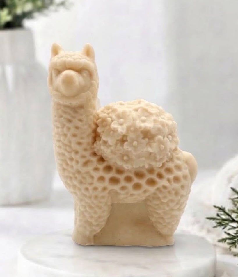 A close-up product photograph of a delicate, yellow French clay llama / alpaca-shaped soap standing on a white marble pedestal. The intricate carving of the soap captures the fluffy fleece texture and a bouquet of small flowers on its back, set against a blurred background of a white vase and green foliage.