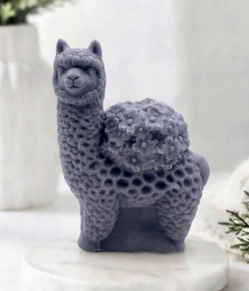 A product photo of a charcoal grey, highly detailed alpaca-shaped soap, standing on a white marble disc. The soap features intricate carvings that depict a fluffy fleece texture and a bouquet of small flowers on its back, placed against a blurred bathroom background with a white vase and green foliage.