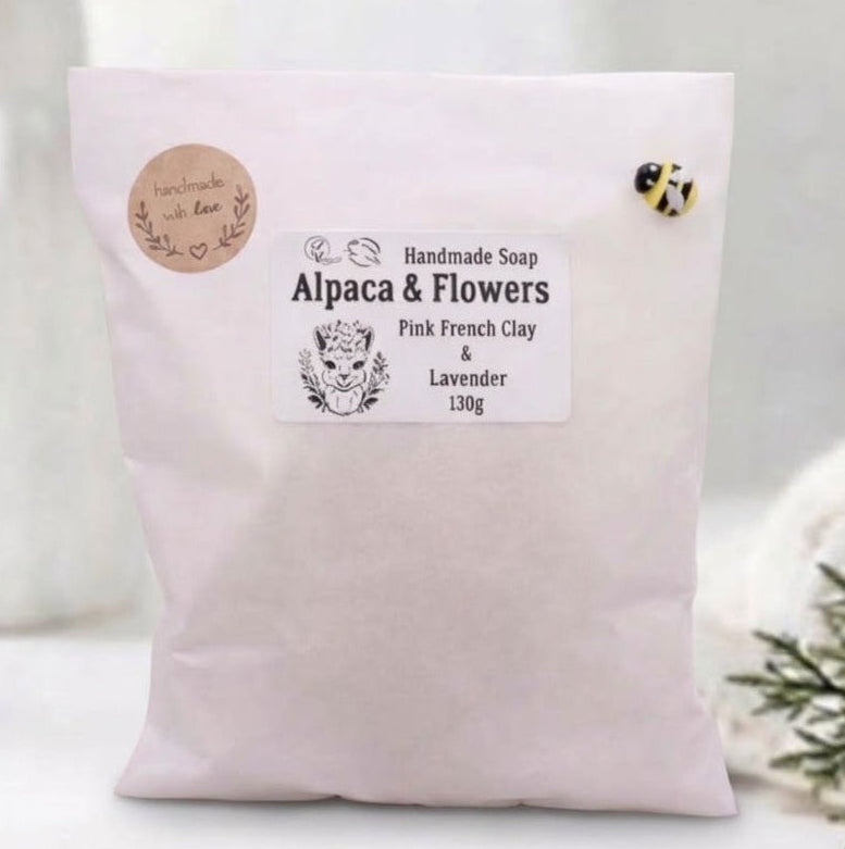 A white paper pouch with product information for "Alpaca & Flowers" handmade soap, placed in a modern bathroom setting with a vase of eucalyptus. The pouch features two small circular brown labels on the left and a small bee-shaped pin on the upper right corner.