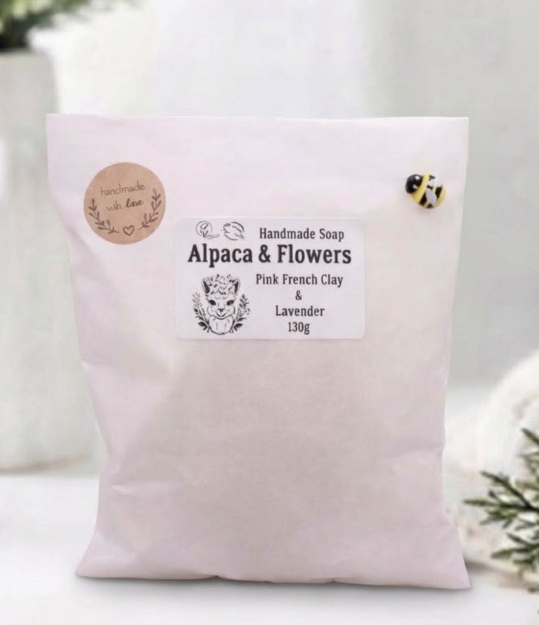 A white paper pouch with product information for "Alpaca & Flowers" handmade soap, placed in a modern bathroom setting with a vase of eucalyptus. The pouch features two small circular brown labels on the left and a small bee-shaped pin on the upper right corner.