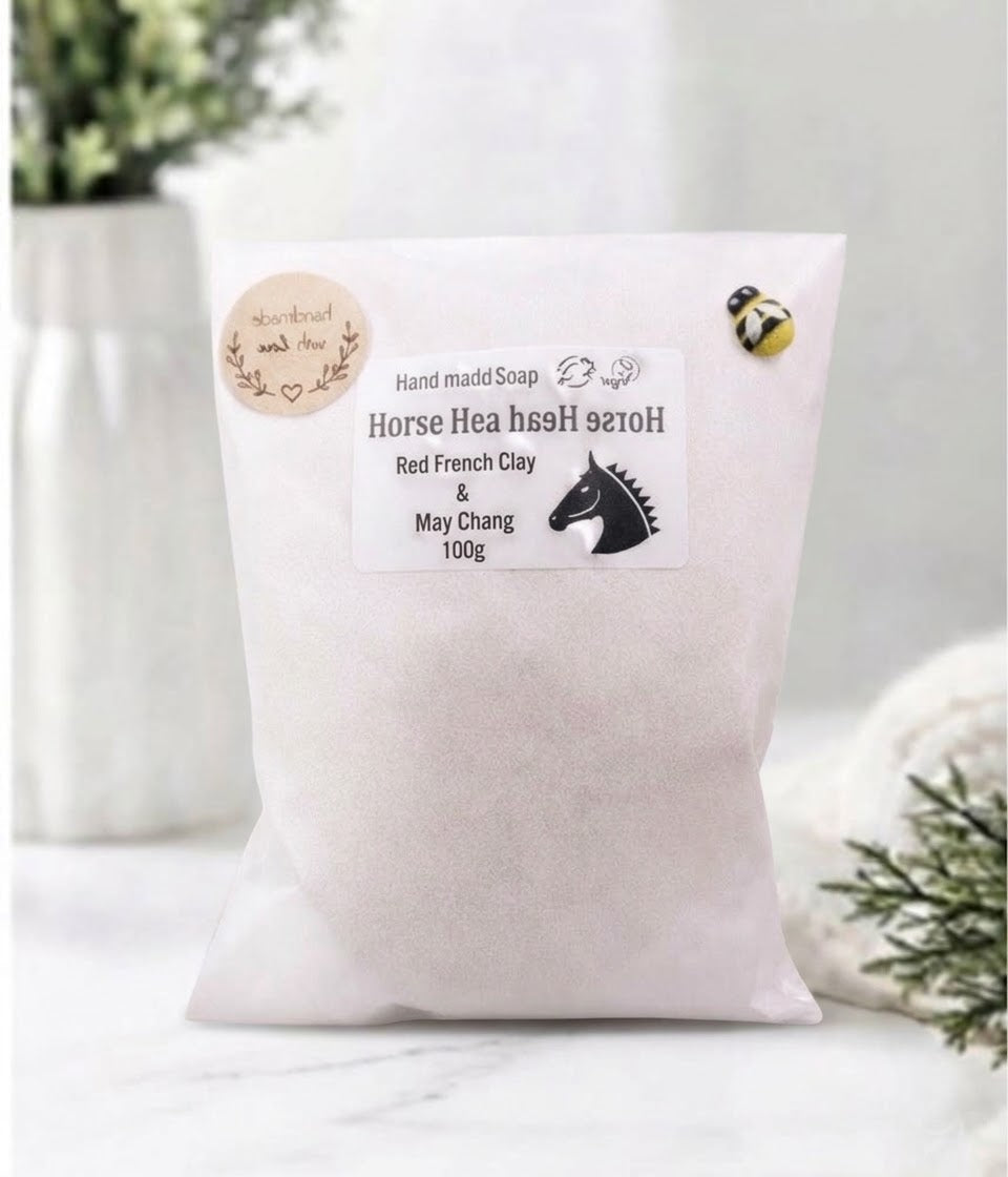 A 100g bag of Horse Hair Soap made from Red French Clay and May Chang clay, featuring a white label with a horse head logo and decorative seals, set against a soft, blurred background with white flowers.
