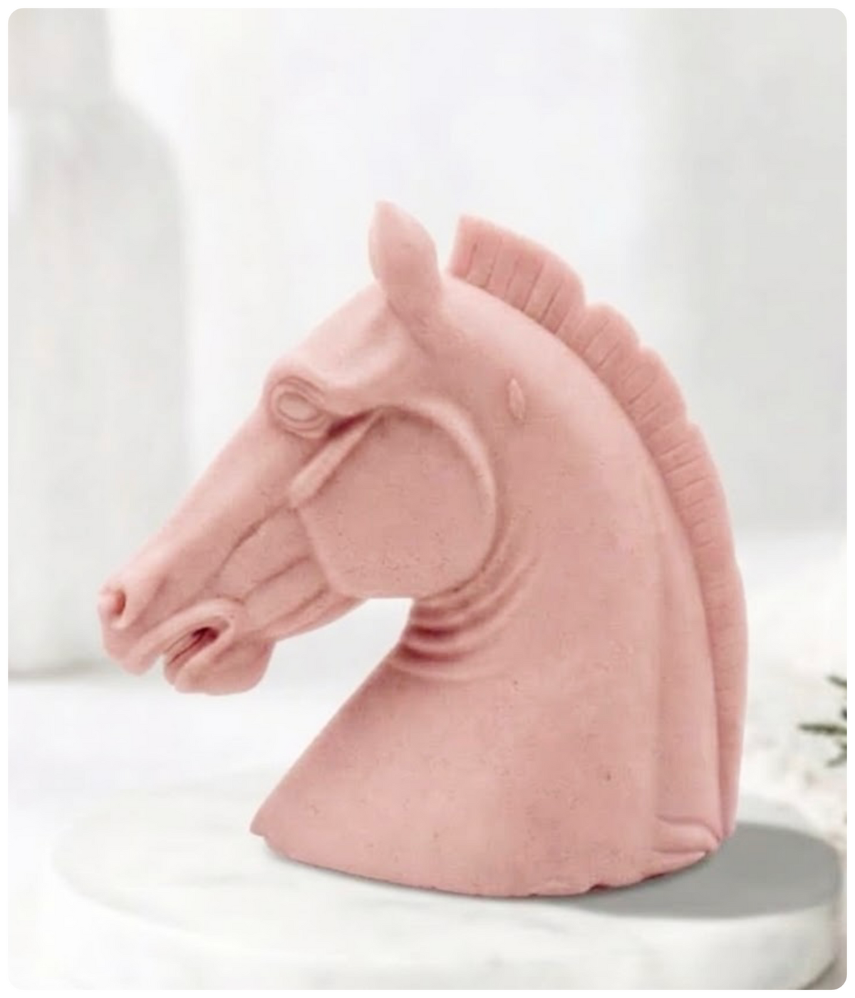 A pink French clay horse head sculptural vegan soap displayed on a white round pedestal against a soft white background.