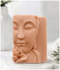 A detailed carved soap bar in a rich terracotta colour, red French clay, depicting the half-face of a serene Buddha holding a lotus flower. It sits on a round marble base in a bright studio setting with blurred decorative items in the background.