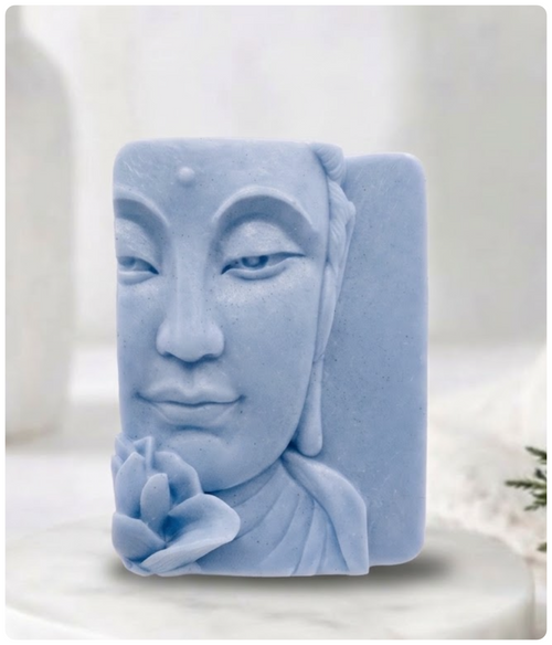 A rectangular bar of light dusty blue artisan soap carved with a detailed relief of a serene Buddha face holding a lotus flower. The soap stands vertically on a round white marble plinth against a softly lit, blurred white background featuring a vase.