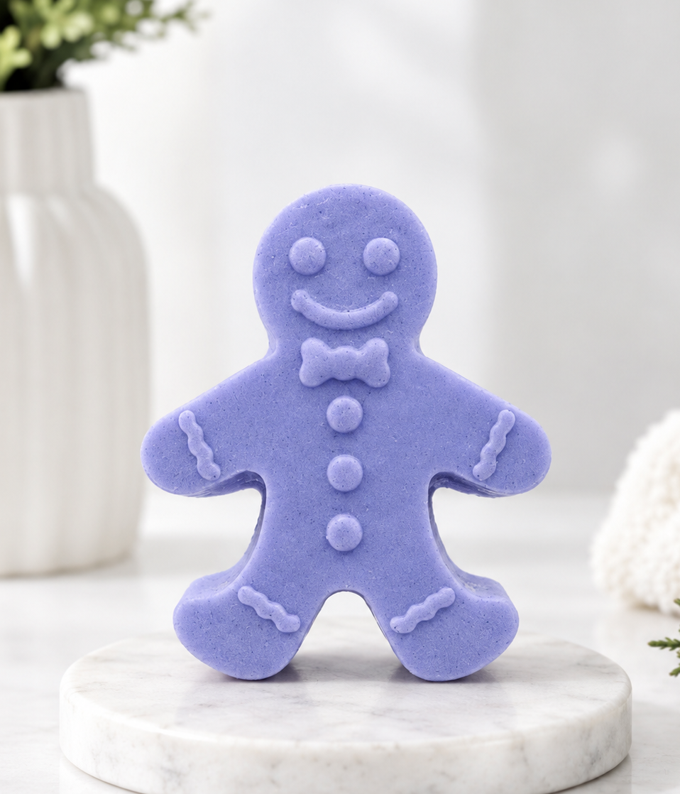 A handmade vegan soap made with indigo powder, featuring a classic gingerbread man with a smiling face, a bow tie, and decorative icing details on his arms and legs, displayed on a miniature wooden easel.