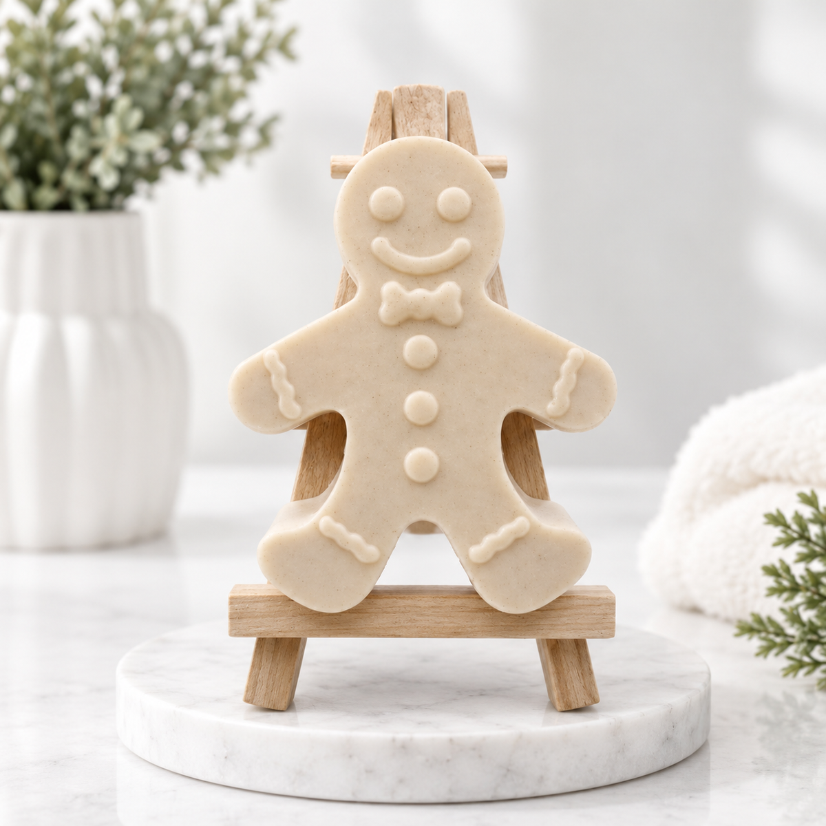 A handmade vegan soap made with yellow French clay, featuring a classic gingerbread man with a smiling face, a bow tie, and decorative icing details on his arms and legs, displayed on a miniature wooden easel.