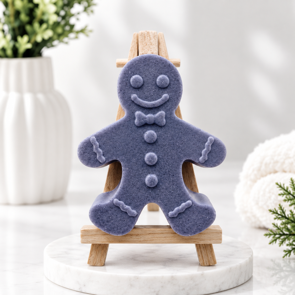 A handmade vegan soap made with activated charcoal, featuring a classic gingerbread man with a smiling face, a bow tie, and decorative icing details on his arms and legs, displayed on a miniature wooden easel.