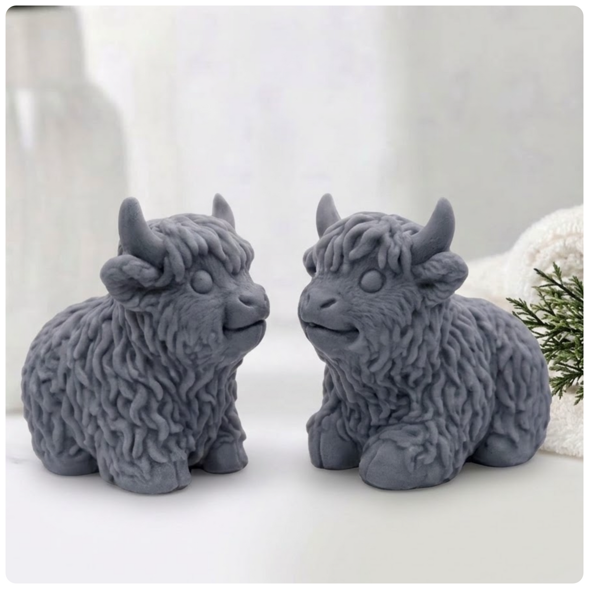Two grey Highland cow vegan soaps made with activated charcoal, sitting side-by-side and facing each other, on a surface with a blurry background of a vase and a towel.