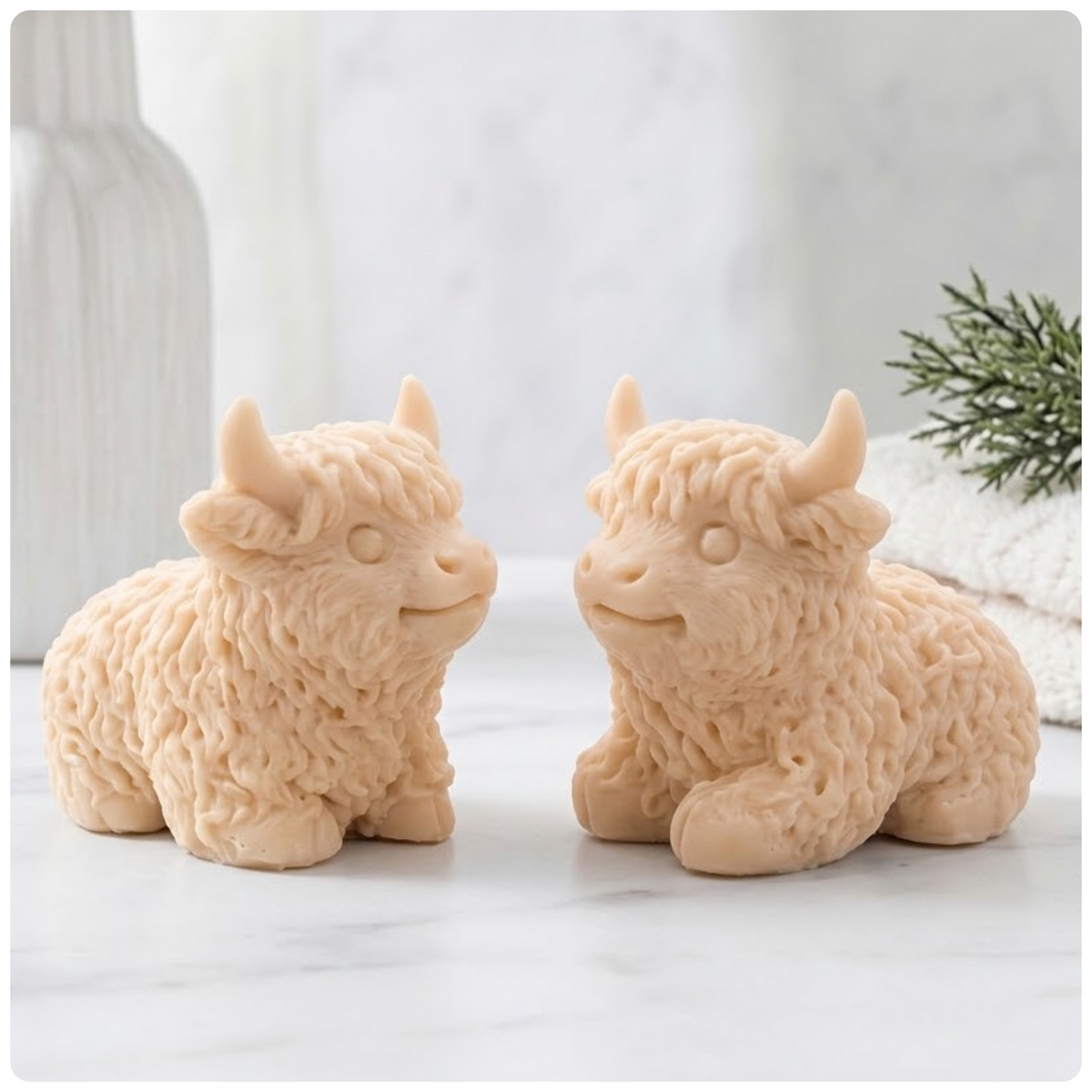 Two natural yellow French clay Highland cow vegan soaps are sitting side-by-side and facing each other, on a surface with a very blurry background.