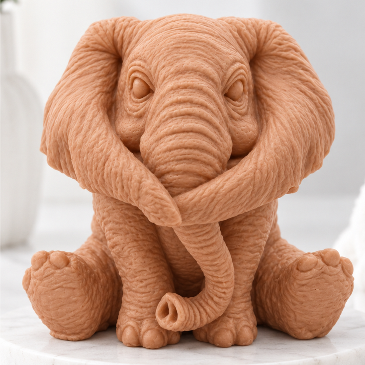 A highly detailed handmade vegan soap gift, made with red French clay, sculpted in the shape of a sitting elephant with realistic wrinkled skin textures and large ears, displayed on a light wooden surface with white flowers in the background.