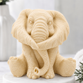 A highly detailed handmade vegan soap gift made with yellow French clay, sculpted in the shape of a sitting elephant with realistic wrinkled skin textures and large ears, displayed on a light wooden surface with white flowers in the background.