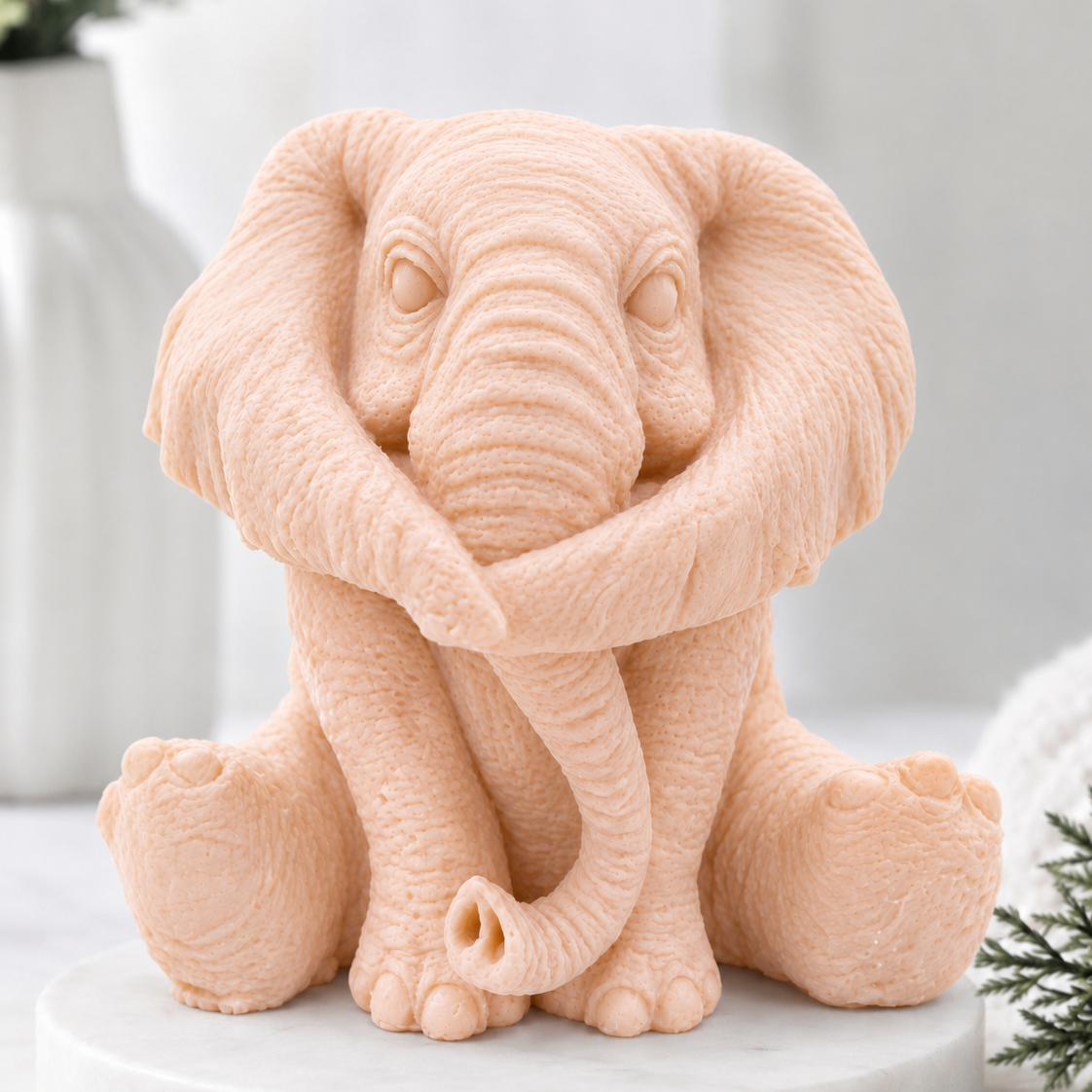A highly detailed handmade vegan soap gift made with pink French clay, sculpted in the shape of a sitting elephant with realistic wrinkled skin textures and large ears, displayed on a light wooden surface with white flowers in the background.