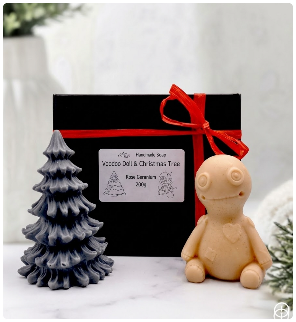 A handmade vegan soap gift set featuring a red French clay voodoo doll soap with stitched details and a activated charcoal grey tiered Christmas tree soap, presented in a black gift box tied with a red raffia ribbon and a small ladybird embellishment.