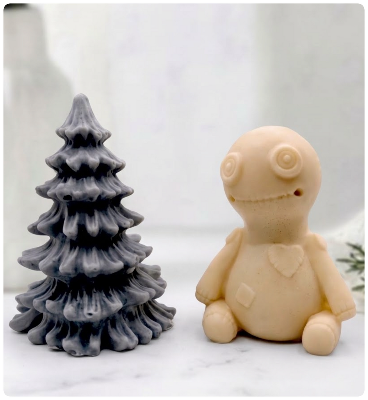 A handmade vegan soap gift set featuring a yellow French clay voodoo doll soap with stitched details and a activated charcoal grey tiered Christmas tree soap, presented in a black gift box tied with a red raffia ribbon and a small ladybird embellishment.