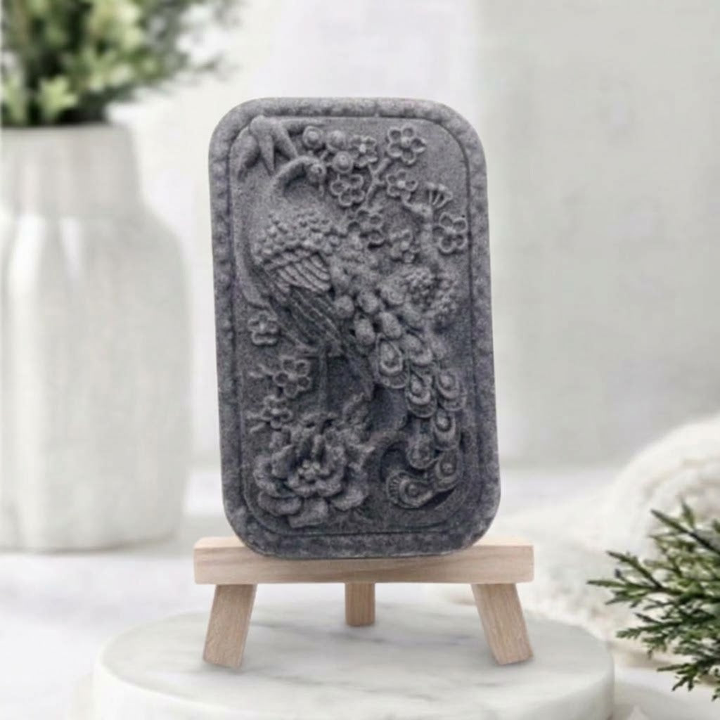 Activated charcoal gray artisanal peacock design vegan soap on a mini easel stand in a spa-like setting.
