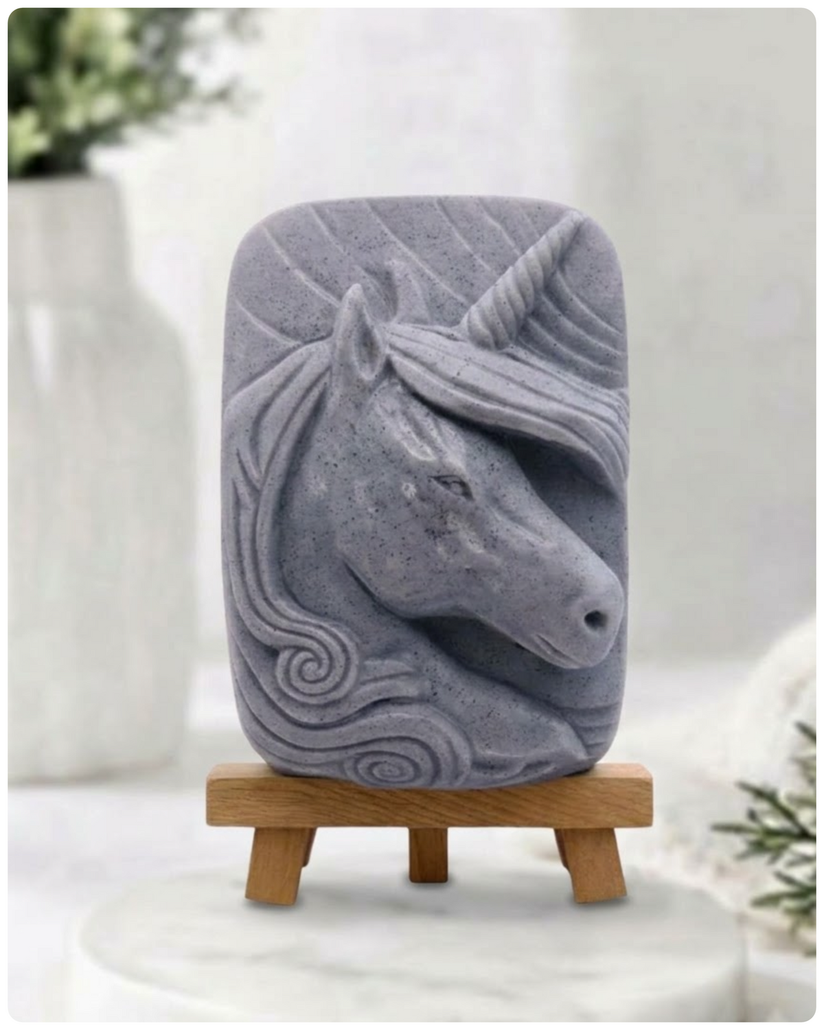 handmade vegan soap featuring a detailed unicorn head relief with a spiralled horn and flowing curly mane, made with activated charcoal and displayed on a small wooden easel. 