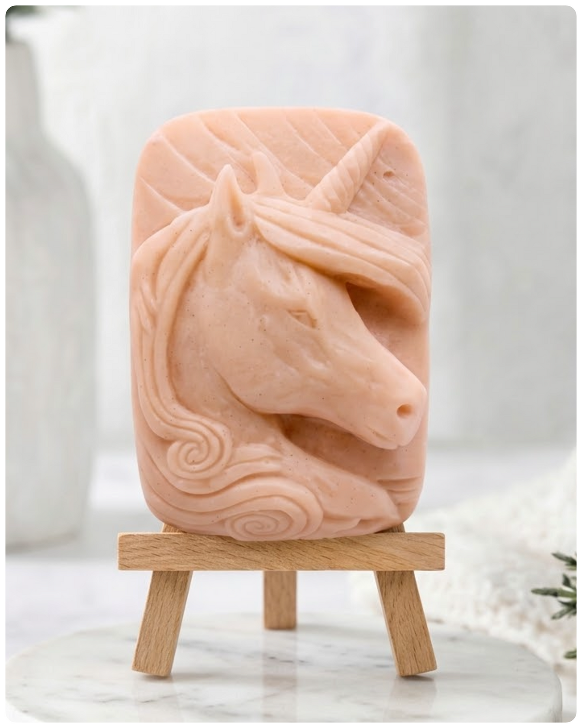 handmade vegan soap featuring a detailed unicorn head relief with a spiralled horn and flowing curly mane, made with pink French clay and displayed on a small wooden easel. 