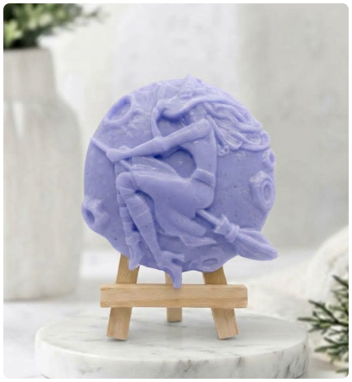 Round, detailed, indigo blue vegan soap block featuring a witch with flowing hair riding a broomstick over a full moon. The soap is displayed upright on a small wooden easel stand.