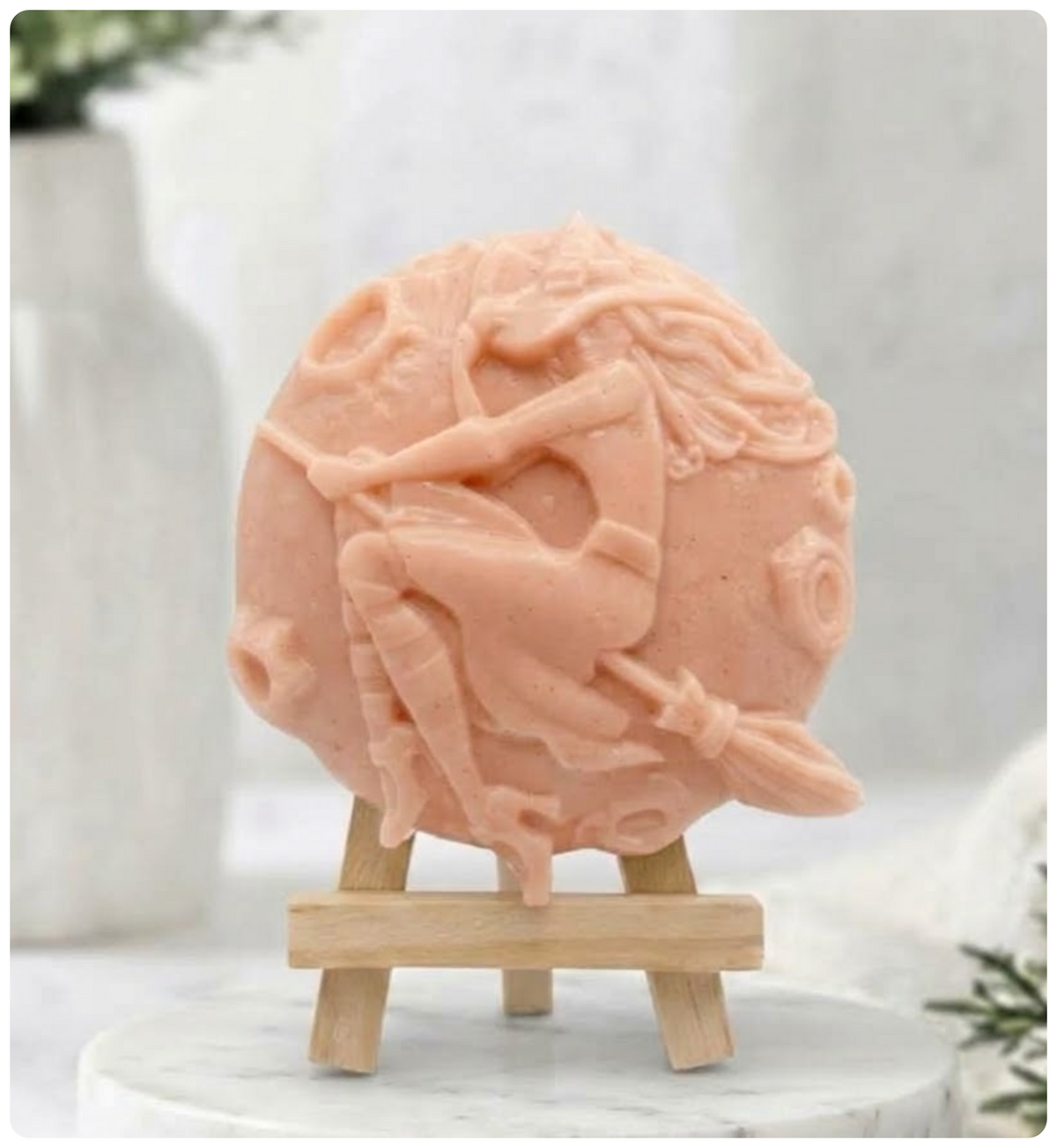 Handmade pink French clay vegan soap carved with a detailed image of a witch riding a broomstick against a full moon. It sits on a miniature wooden display easel.