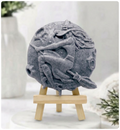 Activated charcoal Grey circular vegan soap carved in intricate detail with a witch on a broomstick flying over a full moon. It is displayed on a miniature wooden stand on a marble plinth.