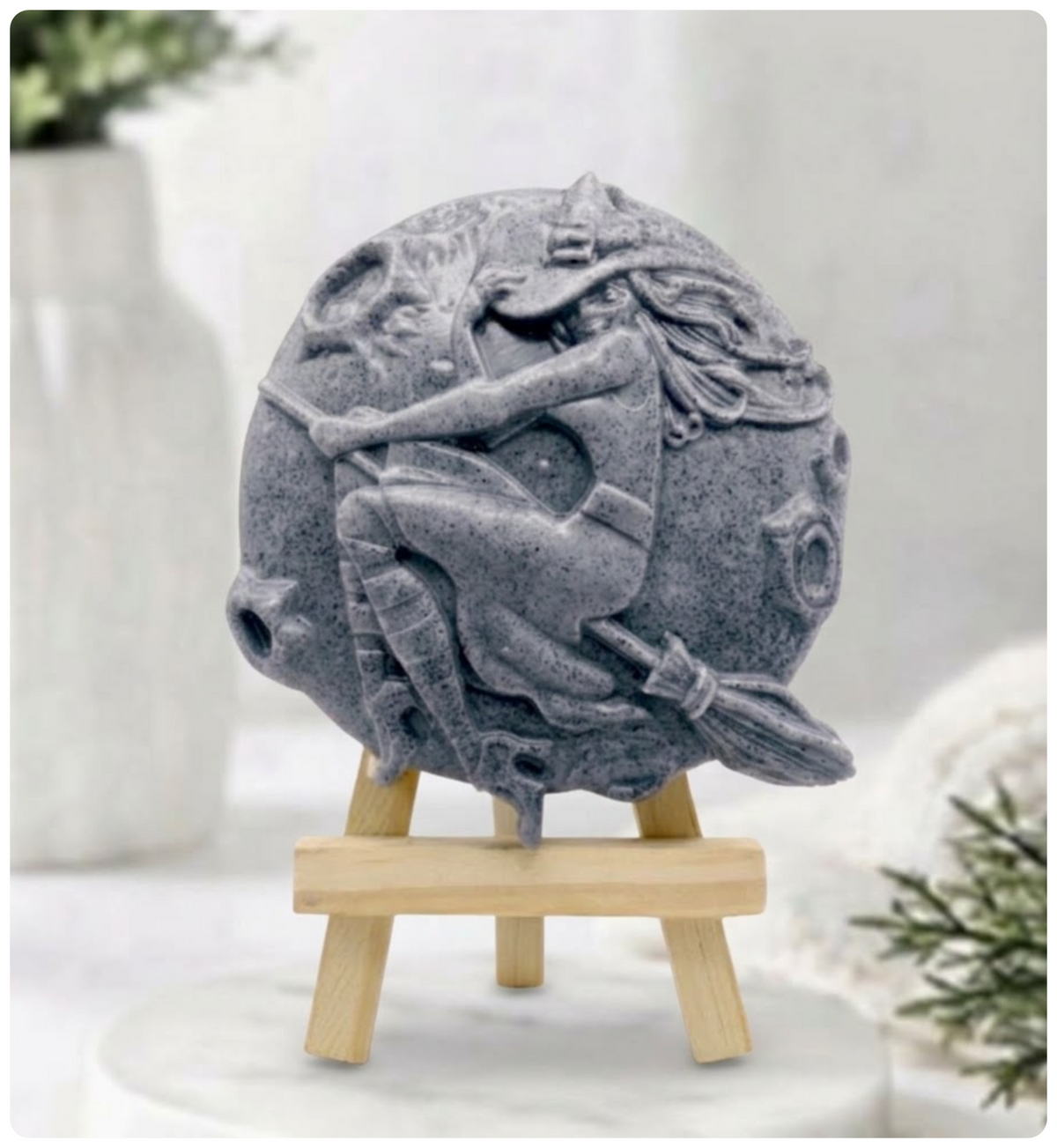 Activated charcoal Grey circular vegan soap carved in intricate detail with a witch on a broomstick flying over a full moon. It is displayed on a miniature wooden stand on a marble plinth.
