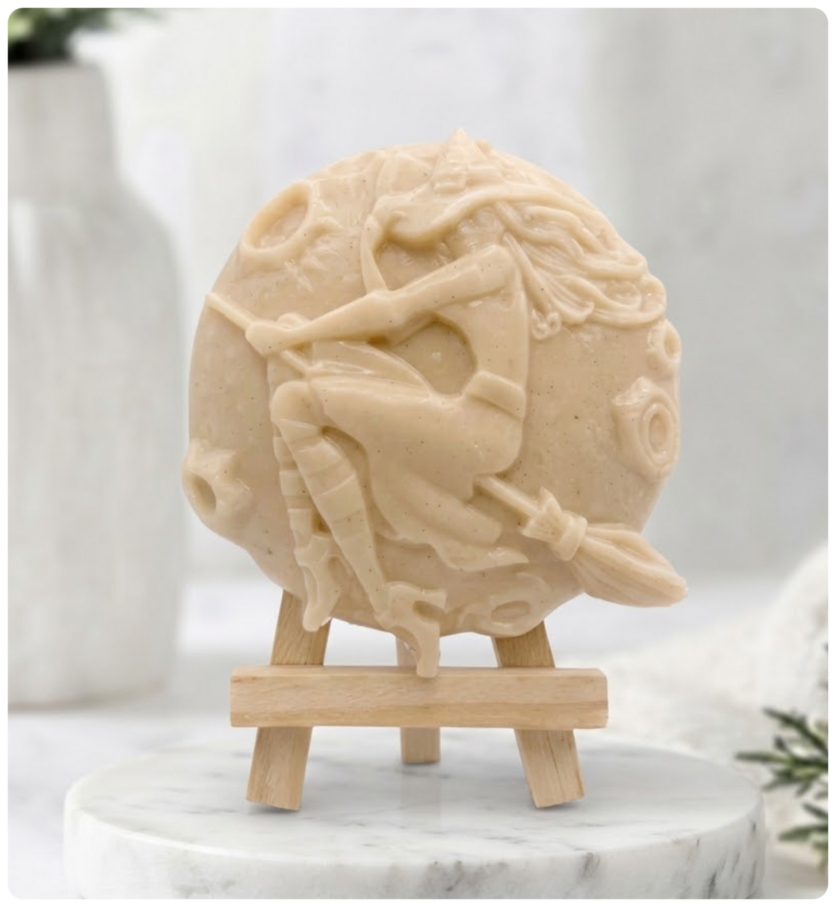 Detailed vegan soap made with yellow French clay in a circular shape. The carving depicts a witch with long hair riding a broomstick over a moon. The product is displayed on a miniature wooden tripod 