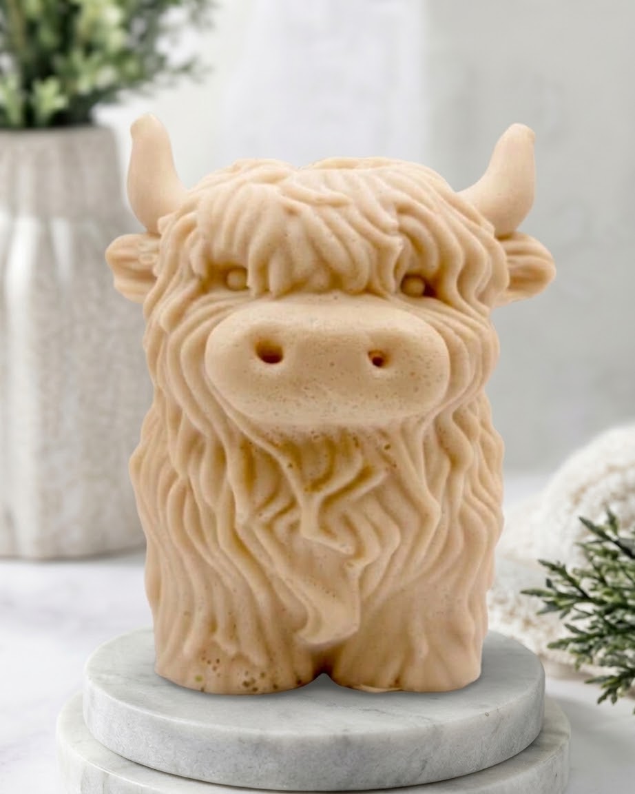 A detailed handmade vegan soap made with yellow French clay, sculpted in the shape of a highland cow with characteristic long, wavy fur, horns, and a friendly face, displayed on a light wooden surface.