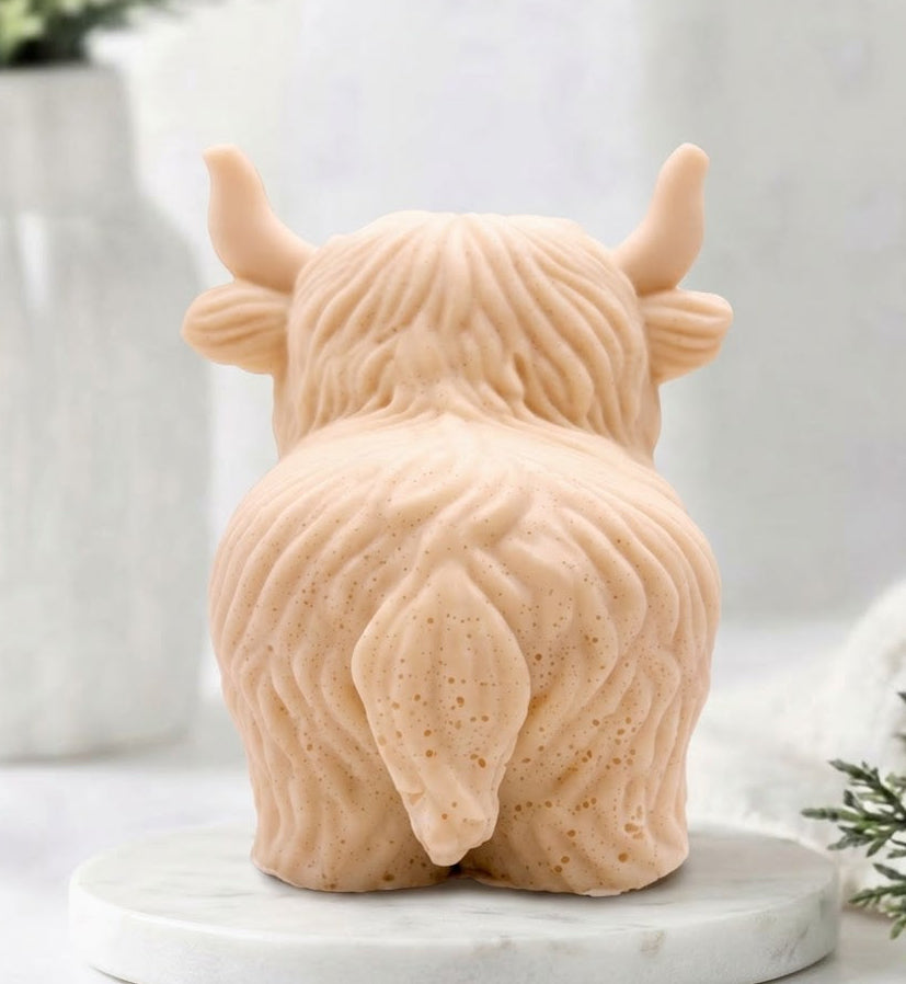 A rear view of a detailed handmade vegan soap, sculpted in the shape of a highland cow with characteristic long, wavy fur, horns, and a friendly face, displayed on a light wooden surface.