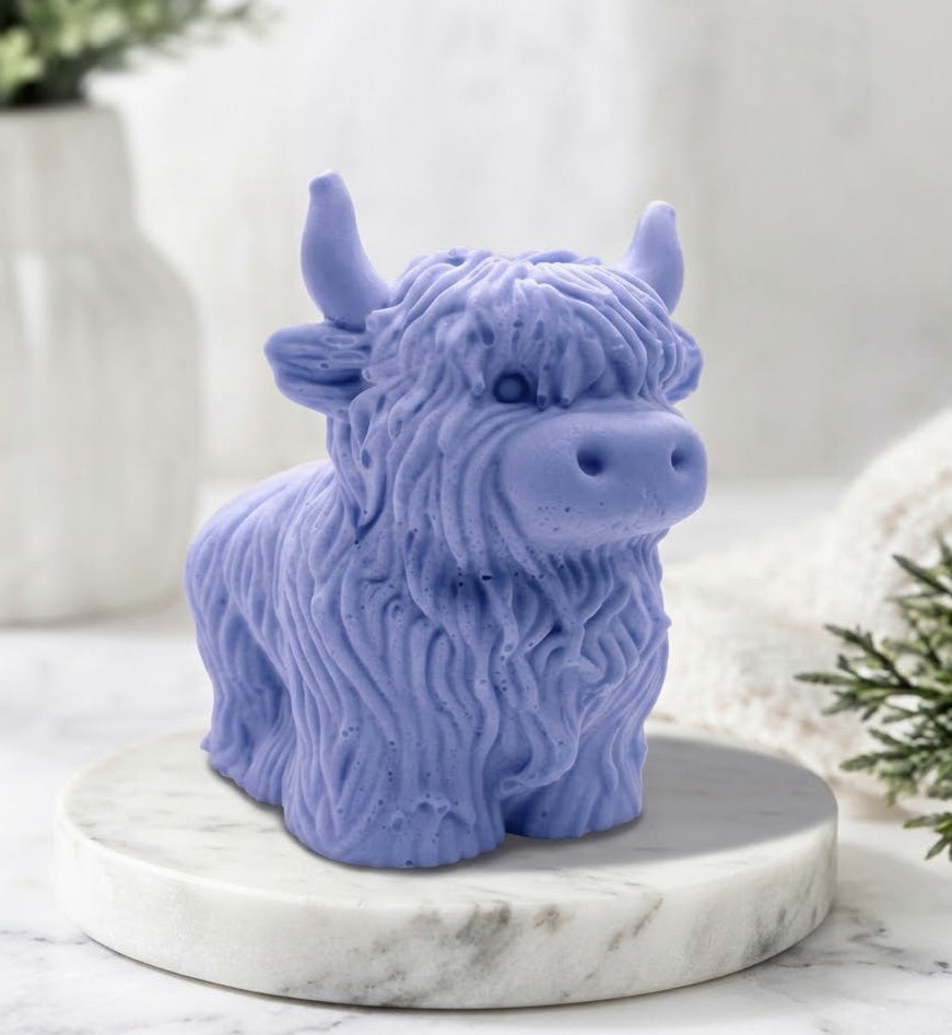 A detailed handmade vegan soap made with indigo powder, sculpted in the shape of a highland cow with characteristic long, wavy fur, horns, and a friendly face, displayed on a light wooden surface.