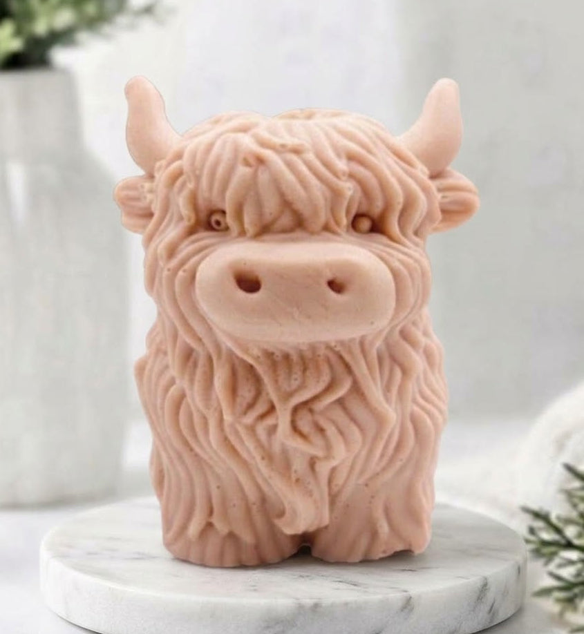 A detailed handmade vegan soap made with pink French clay, sculpted in the shape of a highland cow with characteristic long, wavy fur, horns, and a friendly face, displayed on a light wooden surface.
