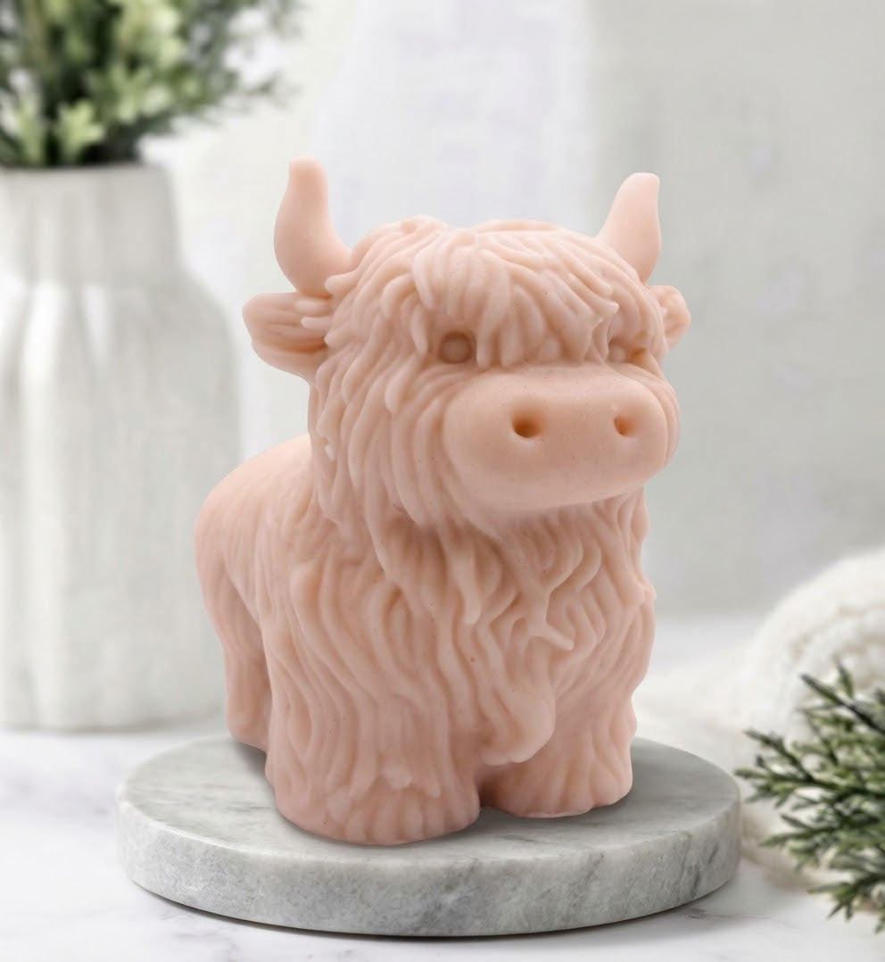 A side view of a detailed handmade vegan soap, sculpted in the shape of a highland cow with characteristic long, wavy fur, horns, and a friendly face, displayed on a light wooden surface.