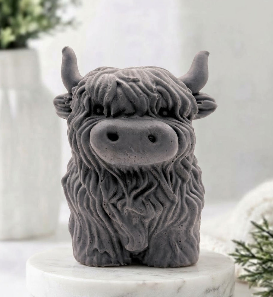 A detailed handmade vegan soap made with activated charcoal, sculpted in the shape of a highland cow with characteristic long, wavy fur, horns, and a friendly face, displayed on a light wooden surface.