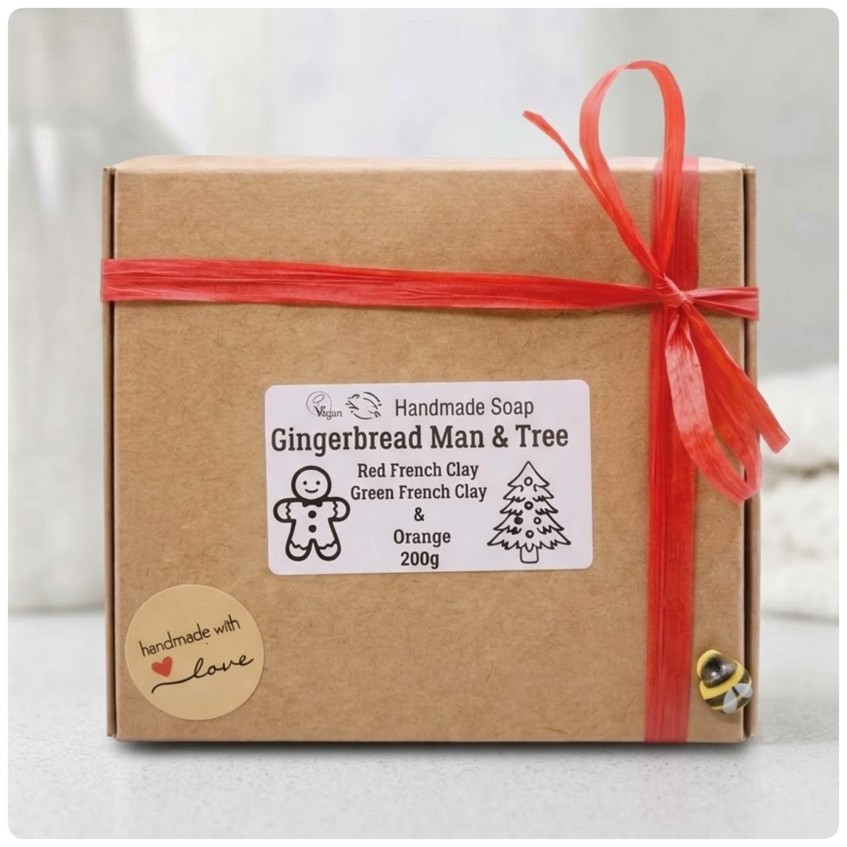 Eco-friendly cardboard gift box containing the handmade vegan Gingerbread Man & Tree soap set, tied with a red raffia bow and decorative bee emblem