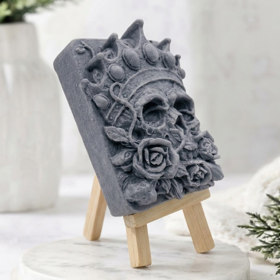 A highly detailed grey carved soap bar featuring a crowned skull with rose flowers on its base, displayed on a small wooden easel over a marble slab in a decorative, spa-like setting.