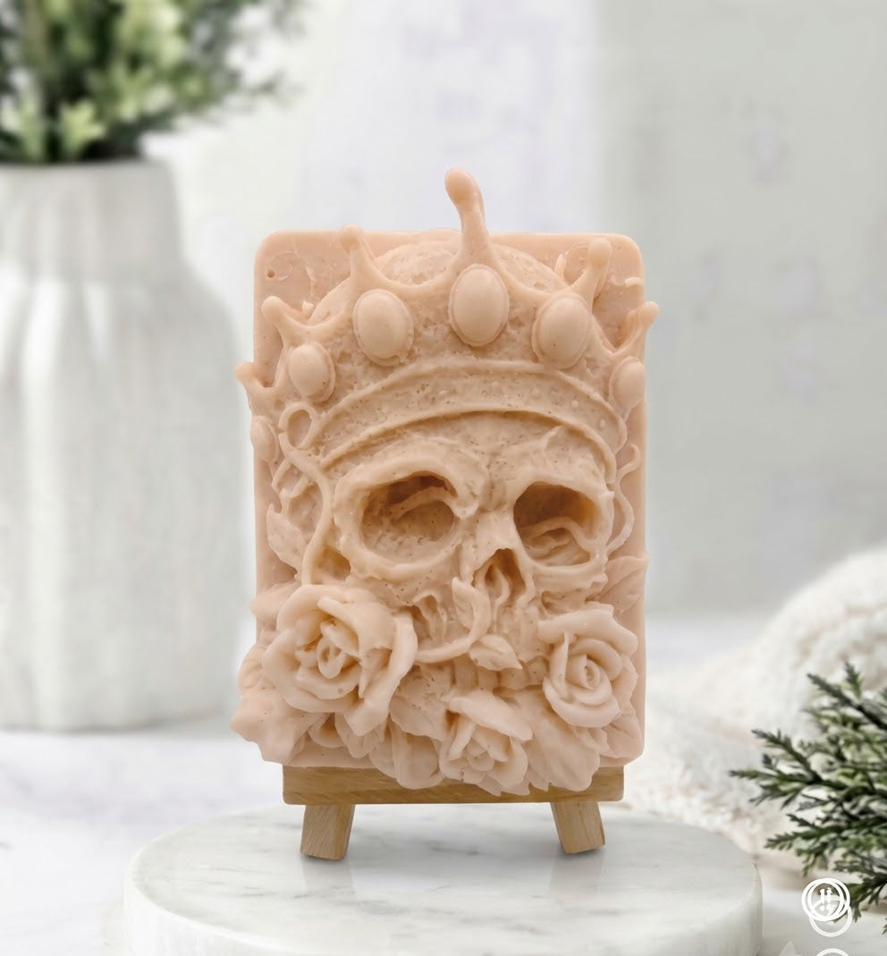 An elaborate close-up image of a pink French clay, finely carved human skull vegan soap with a crown and detailed rose blossoms, resting on a miniature wooden tripod easel over a round marble base in an inviting product presentation.