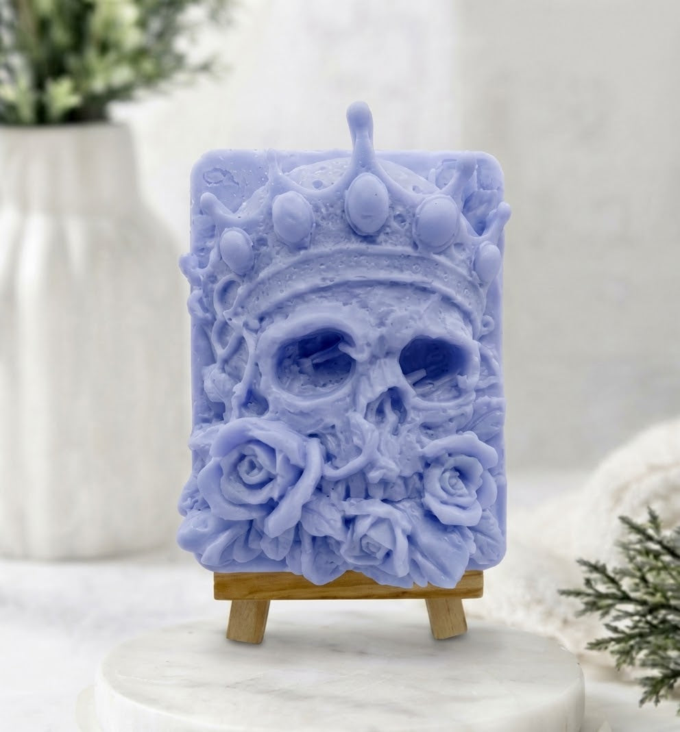A unique blue-coloured soap bar intricately carved into a human skull with a regal crown and detailed rose flowers, presented on a natural wood mini easel in a clean, elegant product shot.