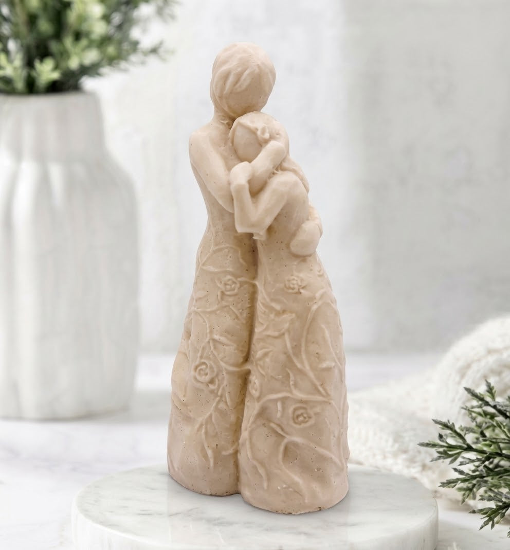 A pale yellow soap sculpture of two figures hugging, standing on a round marble pedestal. The flowing gowns of the figures are detailed with carved rosebuds and vine motifs. A plant in a white vase and a fuzzy white cloth are in the soft-focus background.