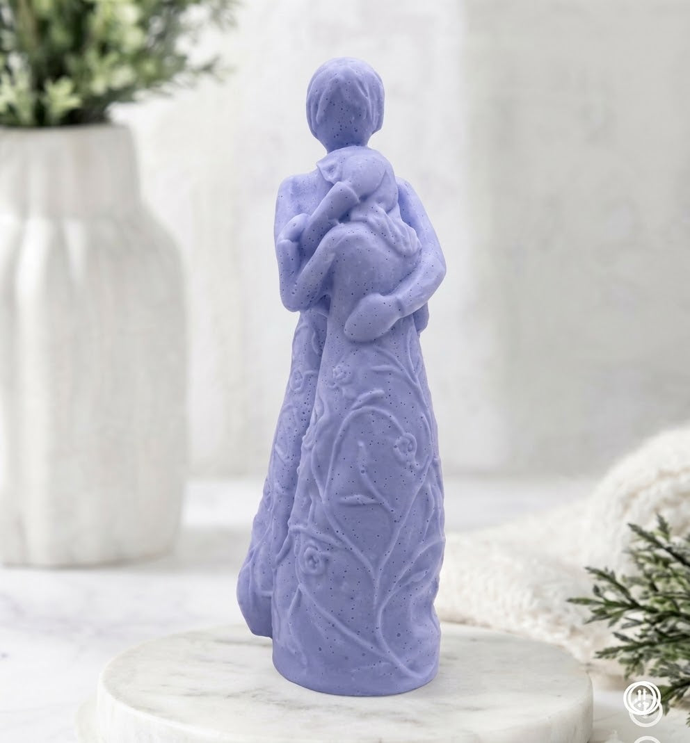 A indigo blue vegan soap carving of two people in a full embrace, standing on a marble pedestal. The figures have no facial features, and their long clothing is adorned with carved flowers and trailing vines. A white vase with green leaves is on the left.