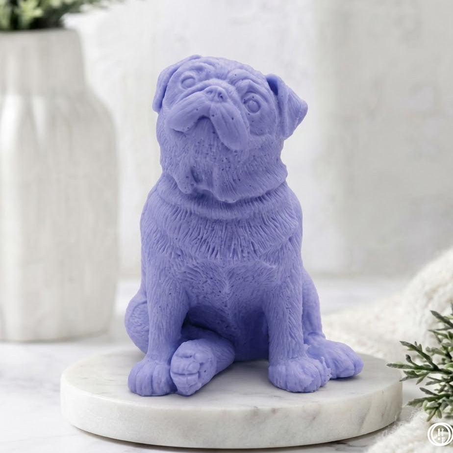 A full-body view from the front of a sitting, sculpted pug dog soap in a vibrant indigo blue colour. Detailed with wrinkles and fur texture, the soap is positioned on a round marble pedestal in a calm spa bathroom setting with a vase of greenery.
