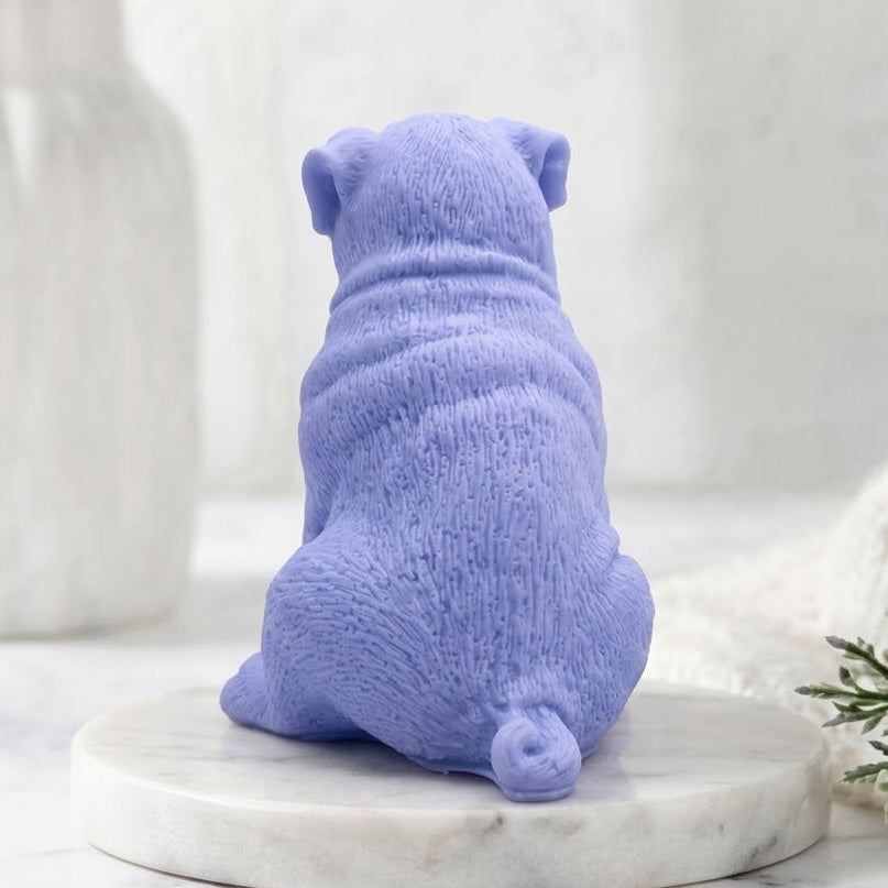 A side view of a sitting, sculpted pug dog soap in a soft blue colour. Detailed with wrinkles and fur texture, the soap is positioned on a round marble pedestal in a calm spa bathroom setting with a vase of greenery.