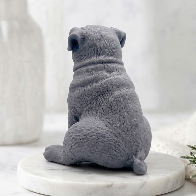 A rear view of a sitting pug-shaped soap, focusing on the back and head of the sculpted gray dog. The soap sits on a round marble pedestal, with a spa environment including a vase and blanket visible in the soft-focus background.