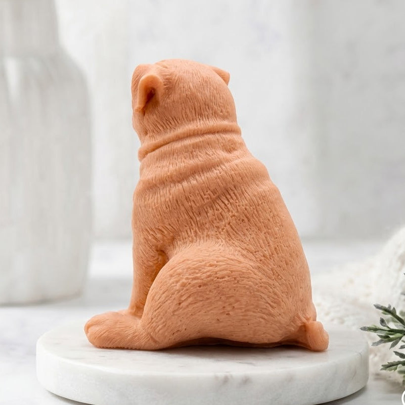 A rear view of the sitting sculpted pug soap, focusing on its back and tail. The soap rests on a round marble pedestal. Soft bathroom elements and greenery are visible in the background.