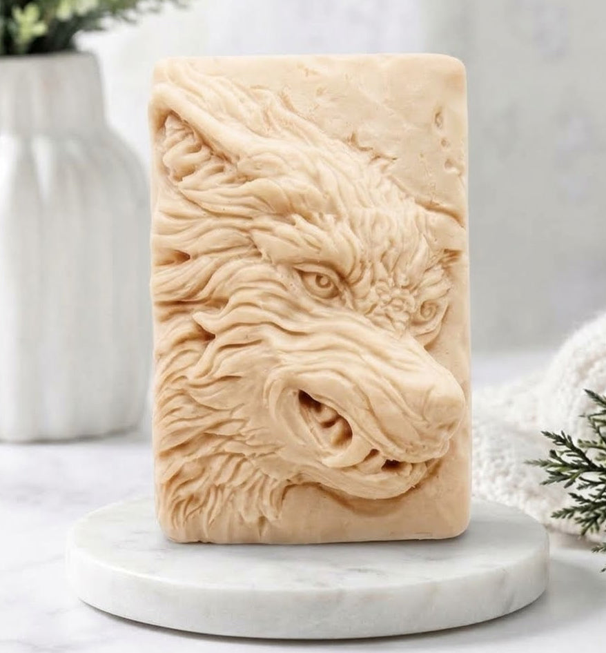 A rectangular yellow French clay soap bar featuring a detailed relief carving of a wolf's head, displayed on a round white marble coaster with a blurred background of a white vase and greenery.