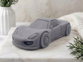 A charcoal grey car-shaped vegan soap styled on a white marble surface with a white textured background and decorative plants.