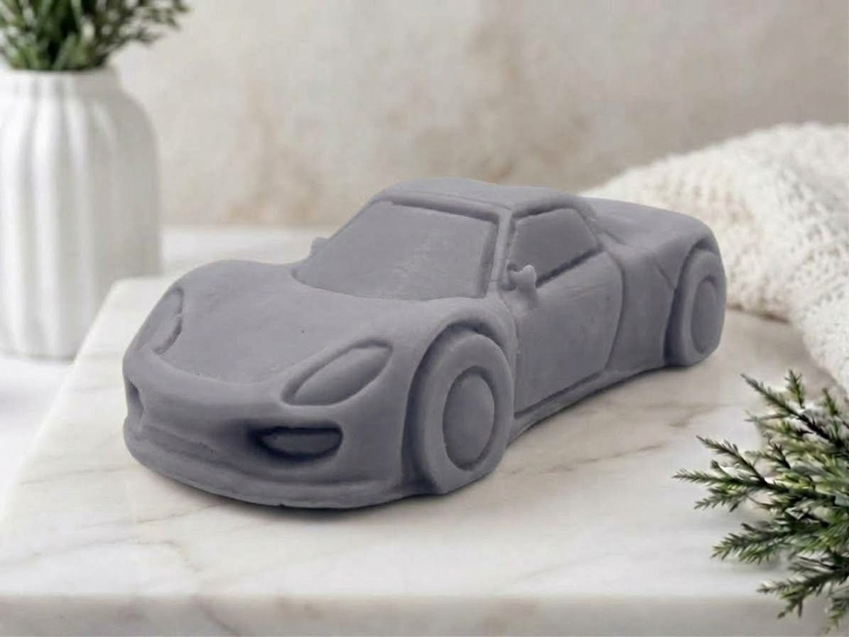 A charcoal grey car-shaped vegan soap styled on a white marble surface with a white textured background and decorative plants.