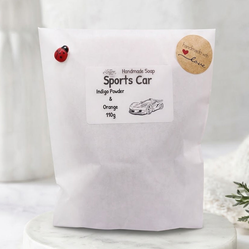 A white stand-up pouch of 'Sports Car' handmade soap indigo powder & orange 190g, displayed on a marble pedestal with decorative plants in the background.