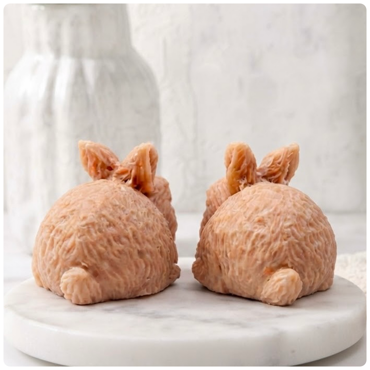 A rear view of two handmade rabbit soaps showing their textured fur and little tails on a marble base.